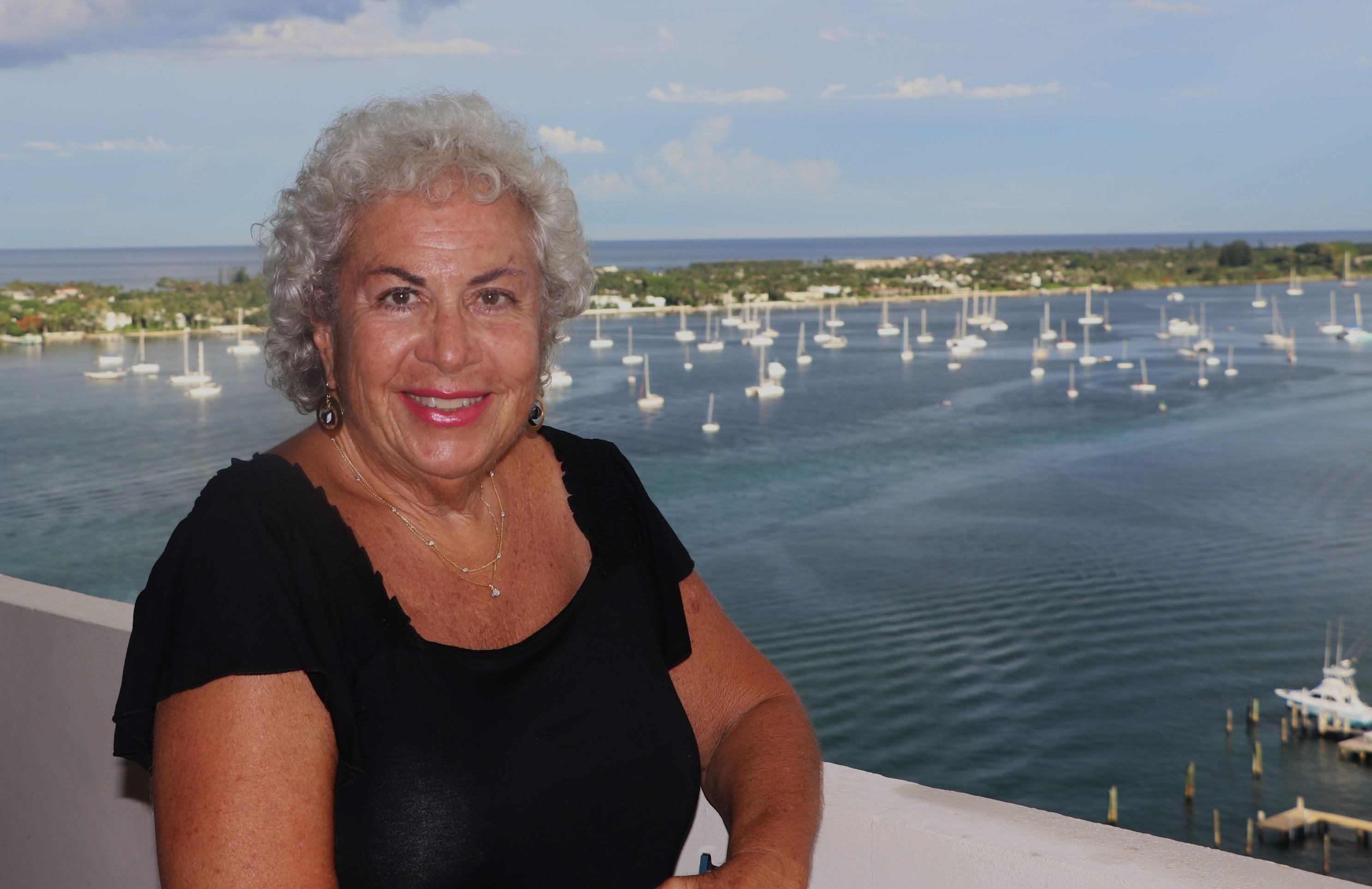 Dina Biblin on a balcony with a backdrop of a harbor with sailboats.